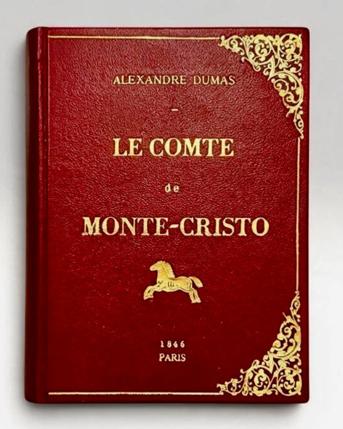 A vintage leather-bound book-clutch of Alexandre Dumas' 'Le Comte de Monte-Cristo' in deep red with gold foil lettering and decorative corner filigree, shown on a neutral light gray background. By M. Design