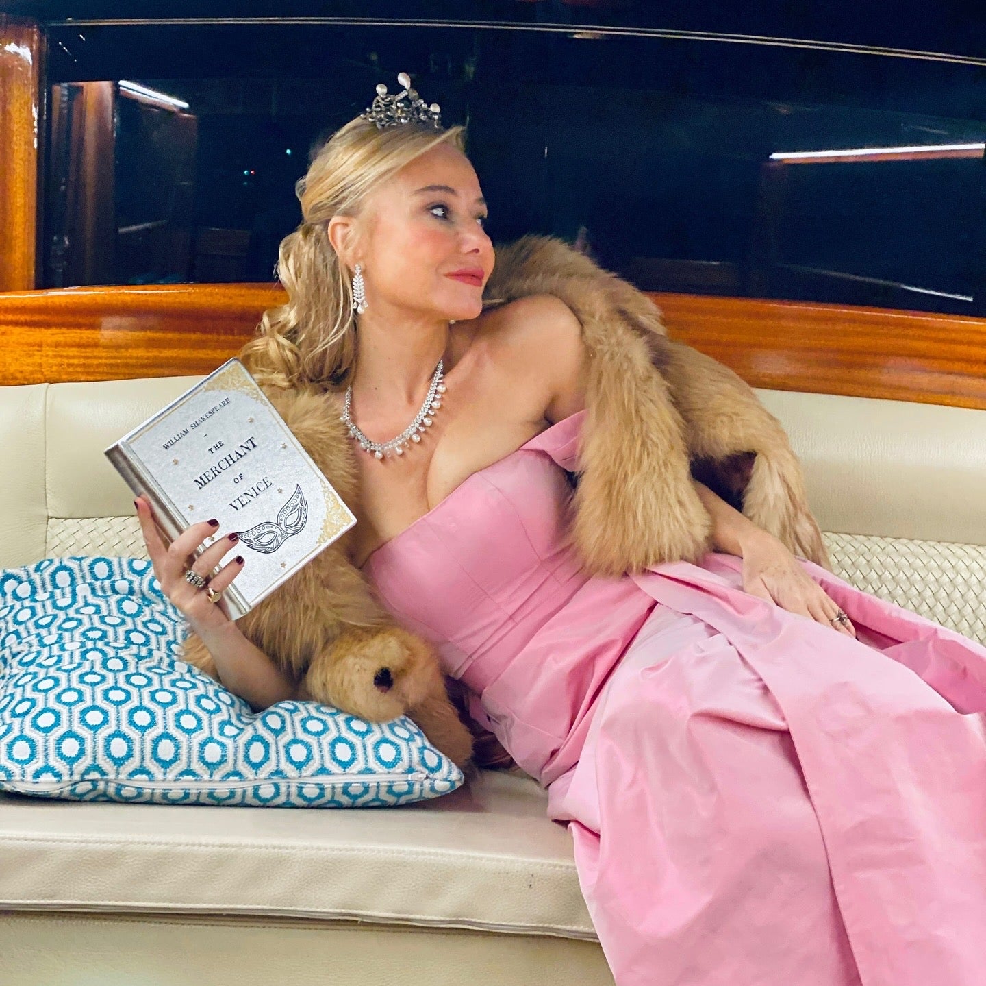 maria marigliano caracciolo in a pink dress with fur coat and tiara, holding a book clutch the merchant of venice, sitting on a boat.