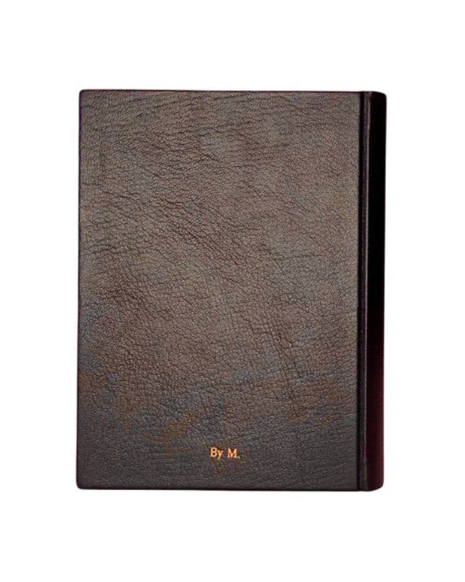 A dark brown textured leather journal or notebook with a black spine and gold 'By M.' monogram on the front, shown against a plain white background.