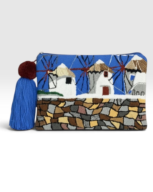 A rectangular woven clutch bag featuring a Mediterranean windmill landscape design with a blue sky, white windmills, and a stone wall. It has a prominent blue tassel with a dark red pom-pom on the left side, set against a plain light gray background.