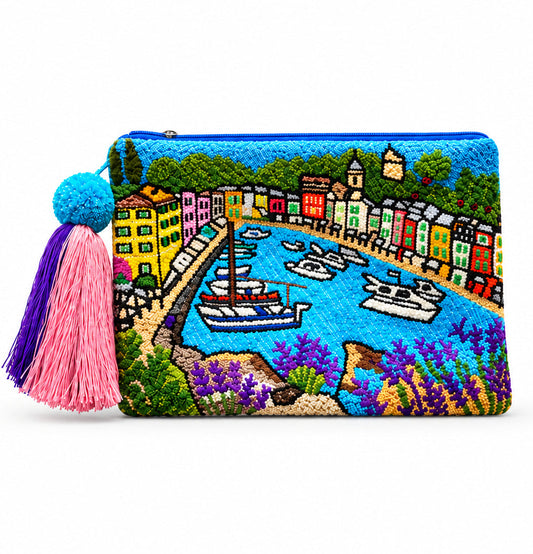 A rectangular embroidered clutch bag featuring a colorful coastal harbor Portofino scene with boats, buildings, and flowers. The bag has a blue tassel zipper closure and is decorated with a large, vibrant tassel in blue, purple, and pink on the left side, set against a plain white background.