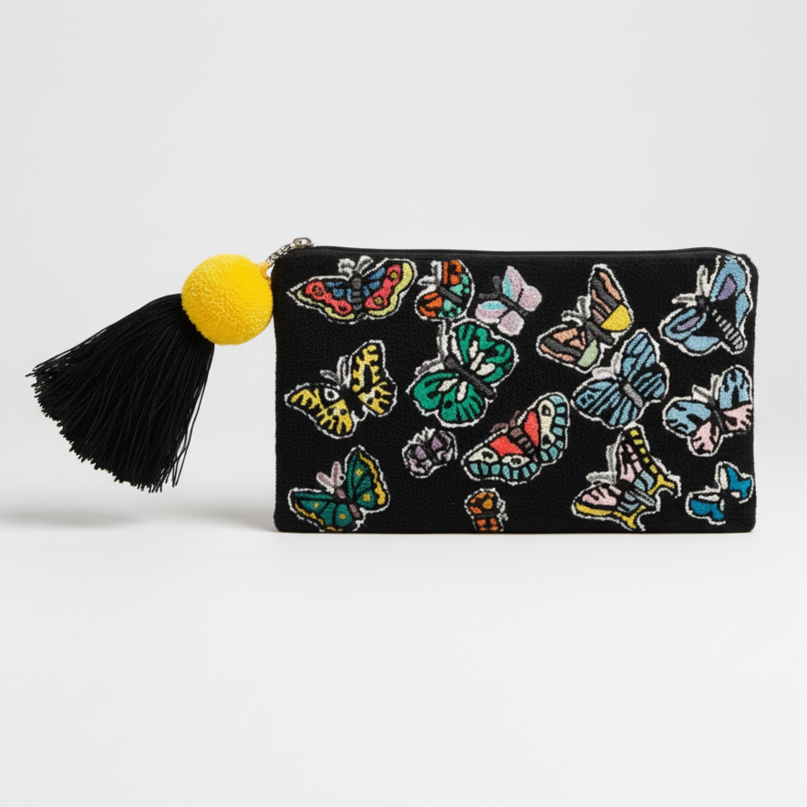 A black zip-around clutch bag decorated with colorful embroidered butterflies, featuring a black tassel with a bright yellow pom-pom on the right side, set against a plain light gray background. By M. design hand woven clutch bag