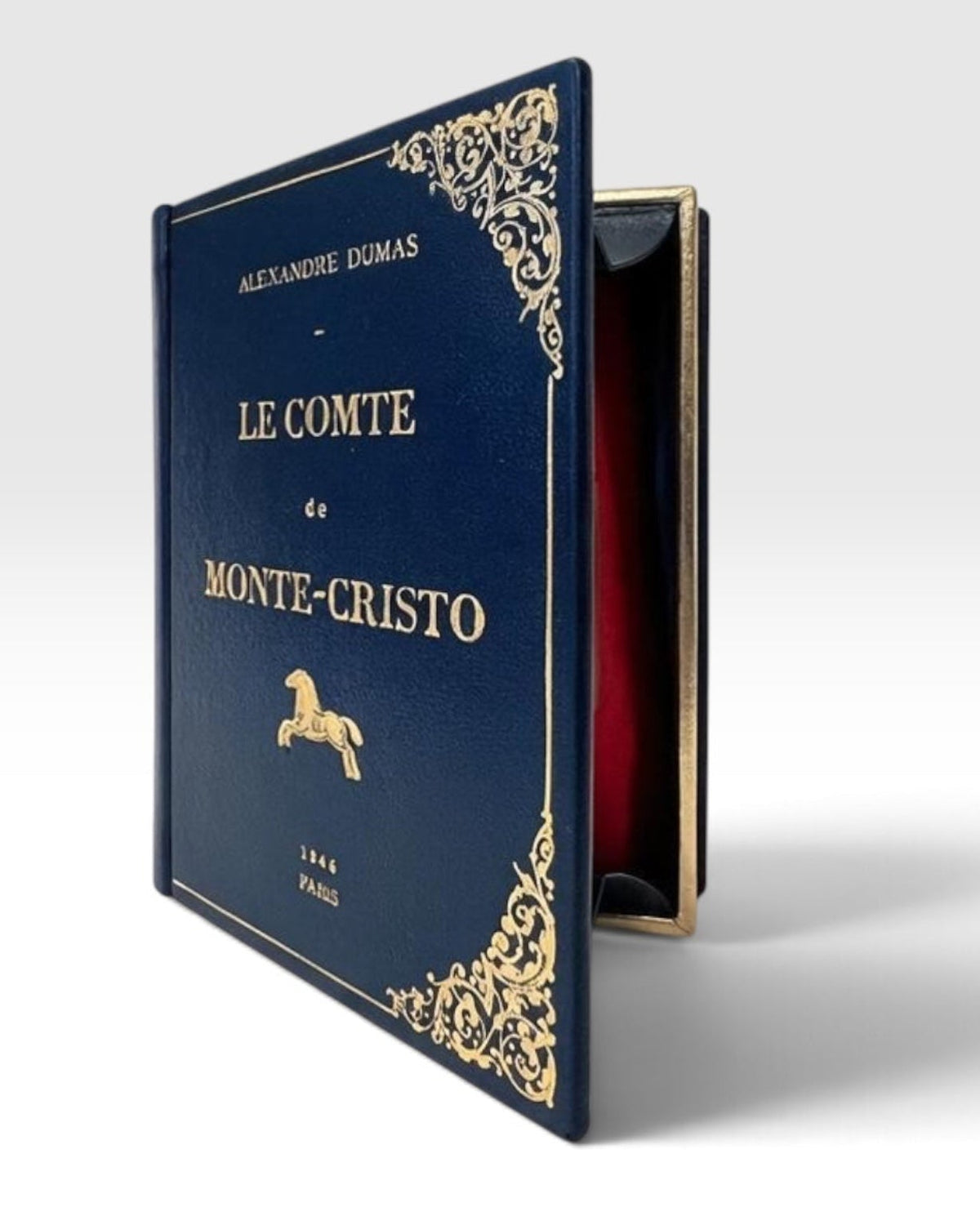 A hardcover edition of Alexandre Dumas' 'Le Comte de Monte-Cristo' book-clutch , featuring a dark navy blue  leather cover with ornate gold filigree corners and gold lettering, standing upright against a light gray background. Maria marigliano caracciolo Design 