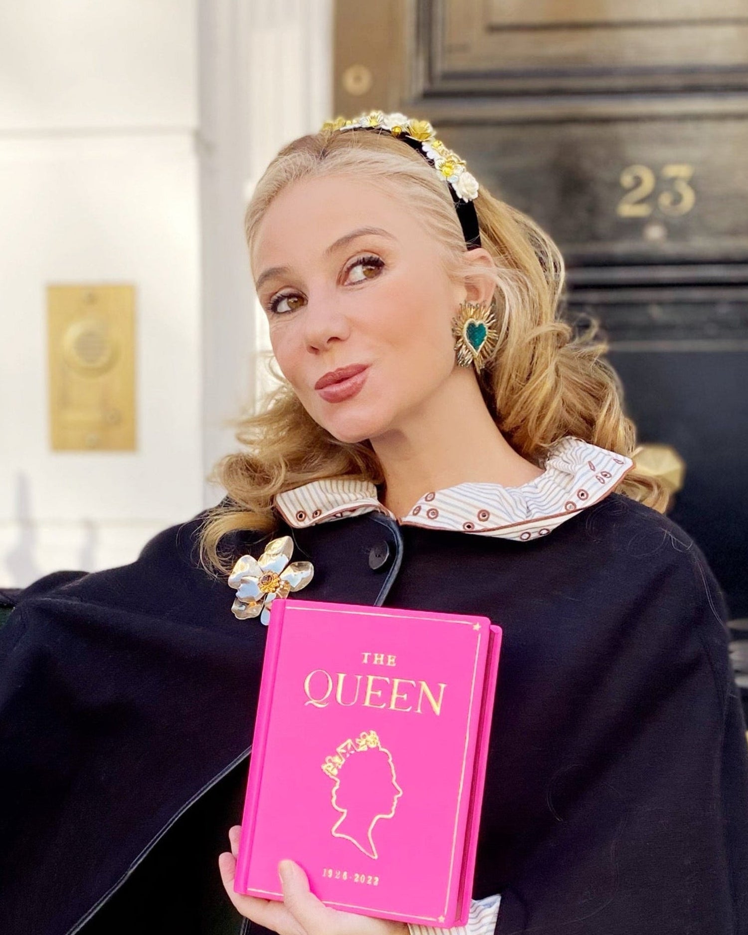 Woman holding a pink book - clutch titled 'The Queen' in front of a building entrance.