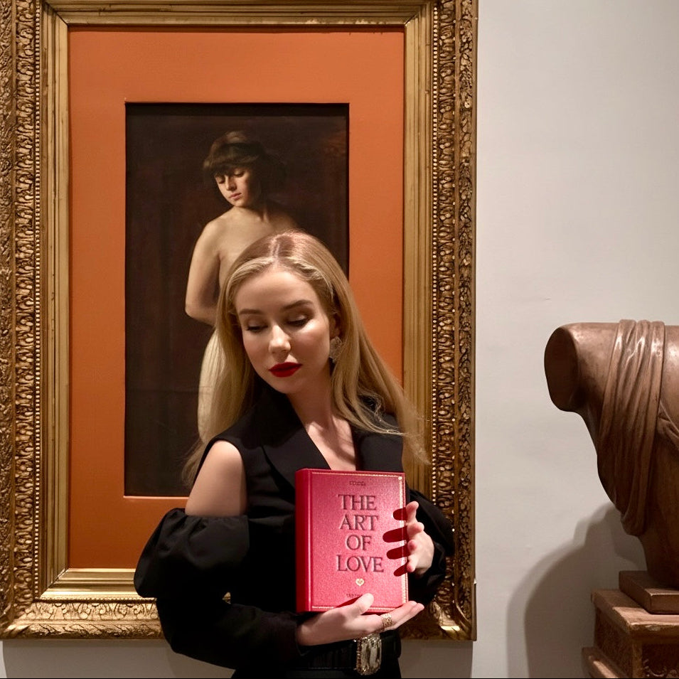 Woman holding a book - clutch titled 'The Art of Love' in front of a framed painting.
